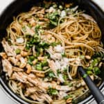 A black pot with spaghetti noodles coated in a sesame cilantro sauce topped with chicken, green onions, and cilantro.