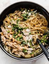 A black pot with spaghetti noodles coated in a sesame cilantro sauce topped with chicken, green onions, and cilantro.