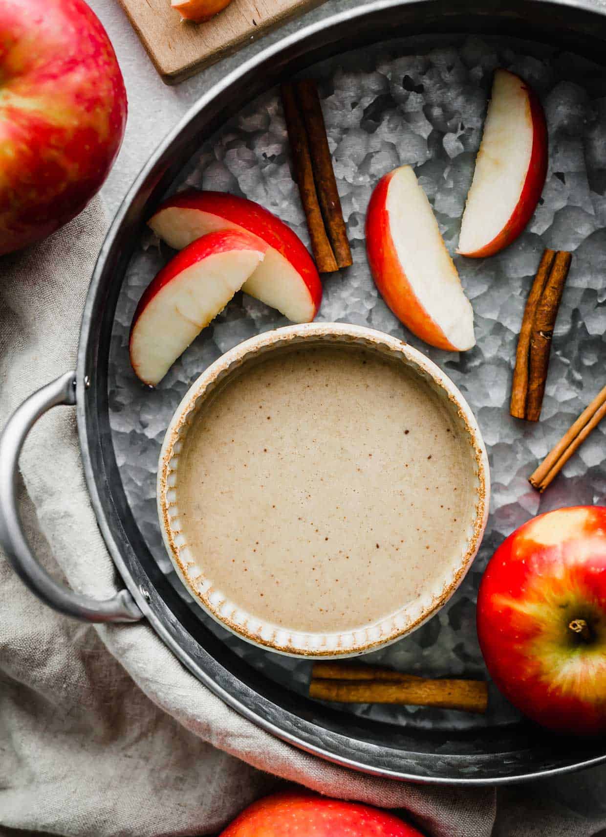 A bowl with Homemade Caramel Apple Dip with cinnamon sticks and sliced red apples in background.