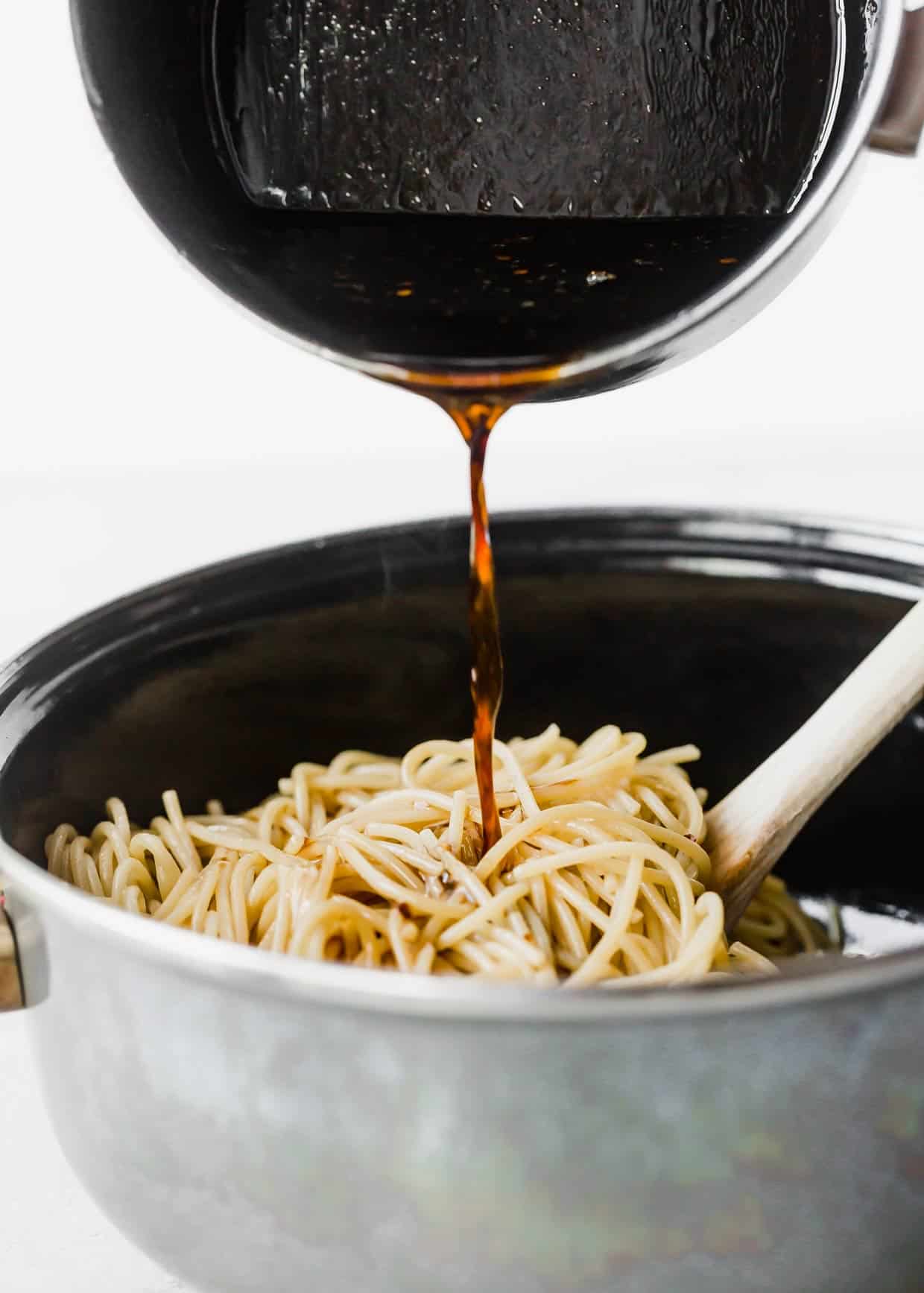 A sesame oil and soy sauce based sauce being poured overtop spaghetti noodles in a black pot.