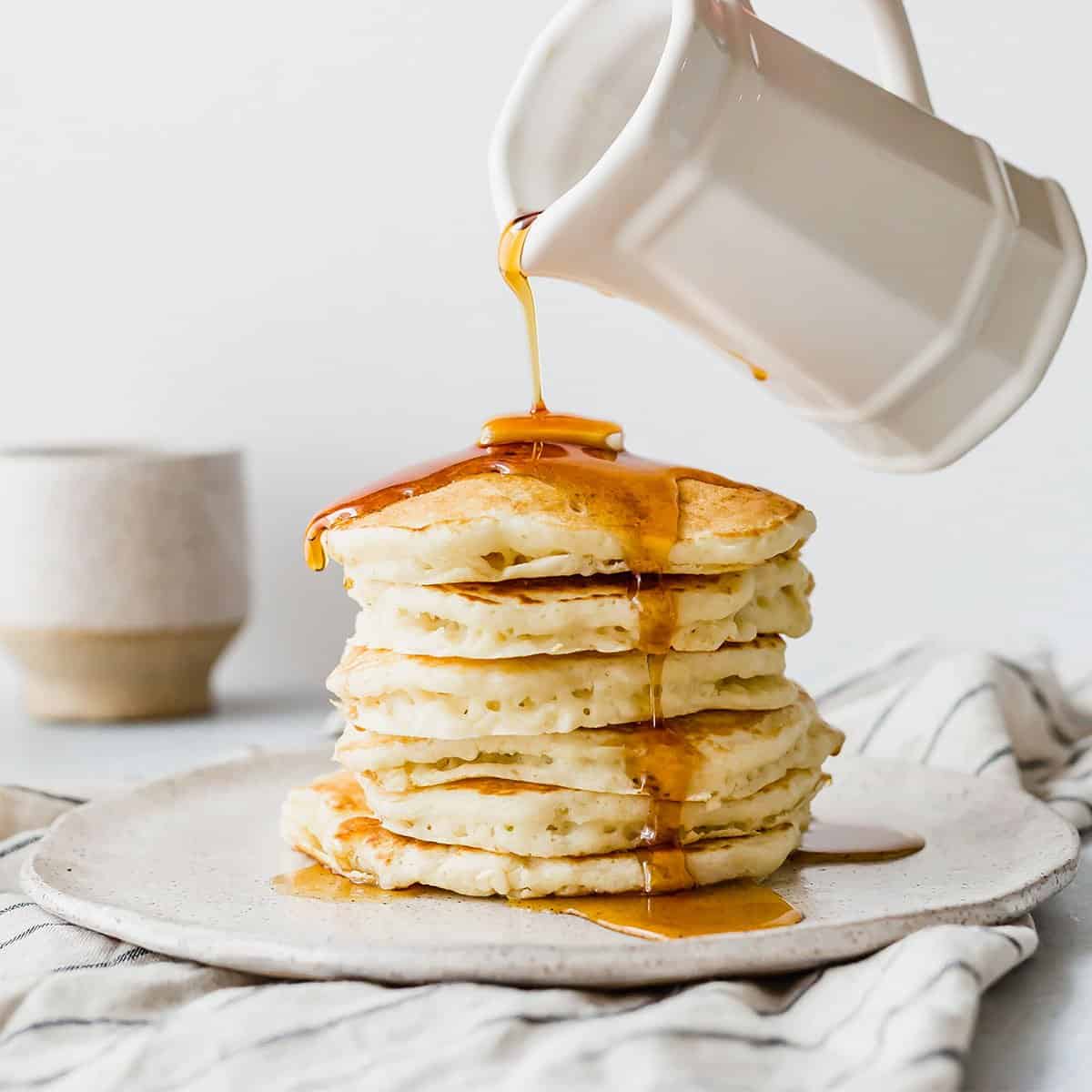 Maple syrup being poured overtop a stack of fluffy buttermilk pancakes on a white plate.