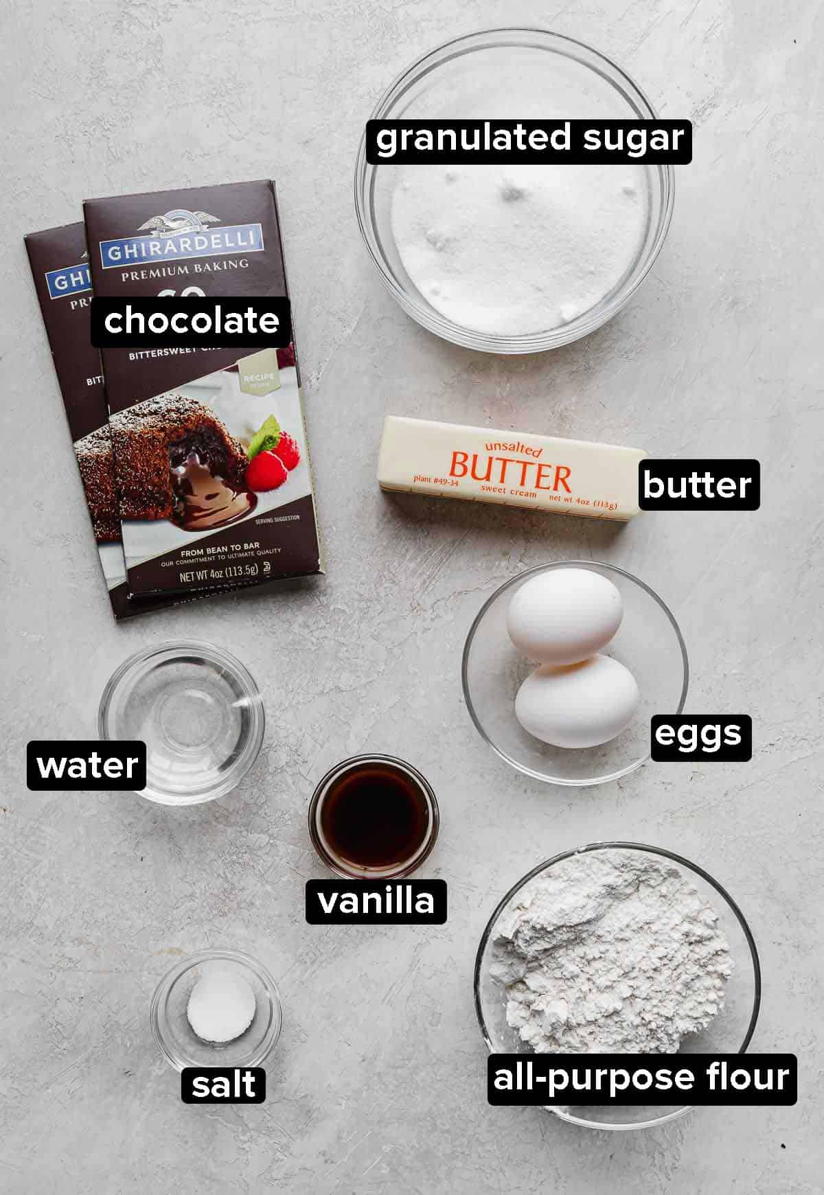 German Chocolate Brownie ingredients: sugar, eggs, butter, flour, vanilla, water, and chocolate bar, on a gray background.