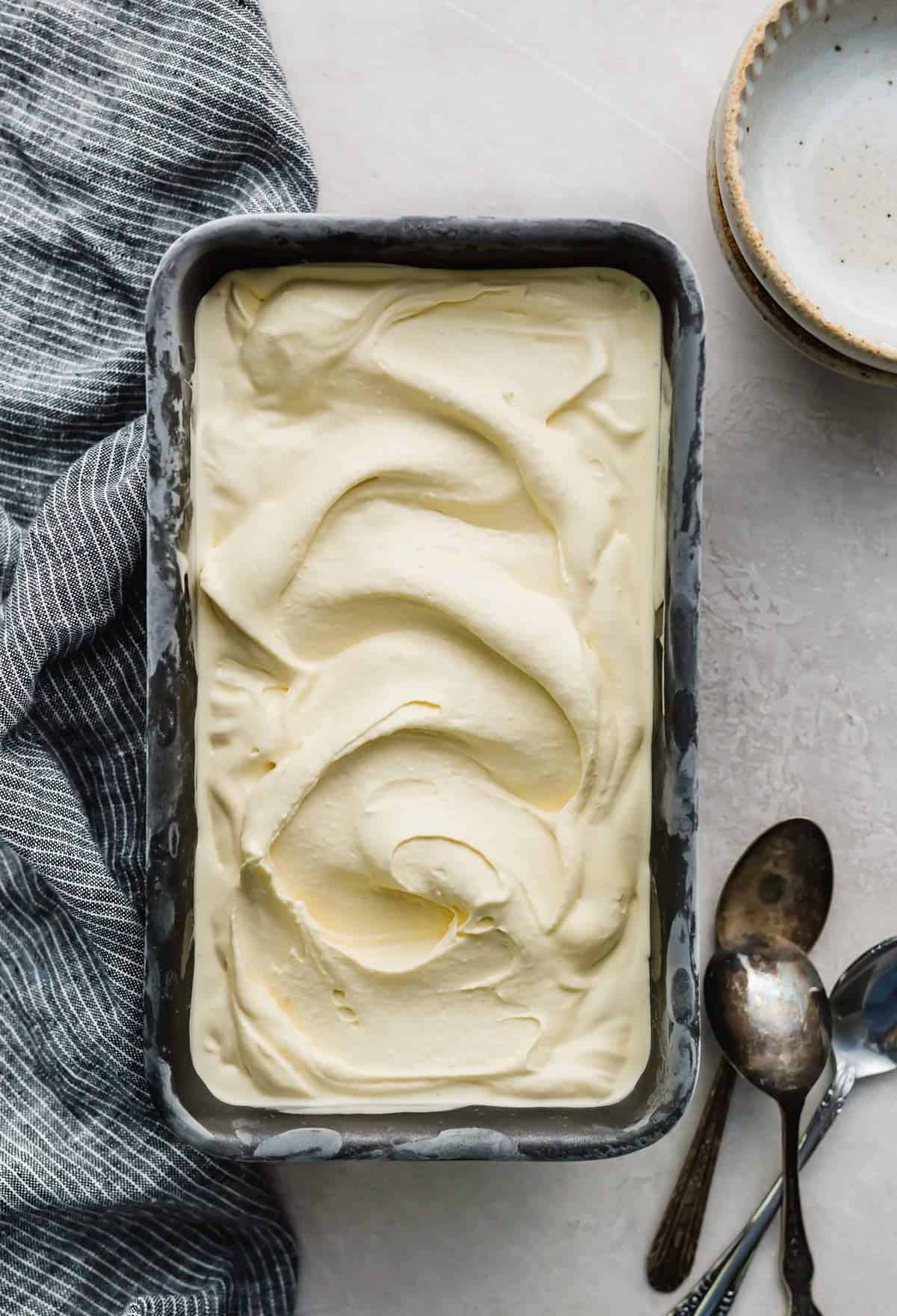 Creamy vanilla ice cream in a dark colored bread pan on a light grey background with spoons next to the pan.
