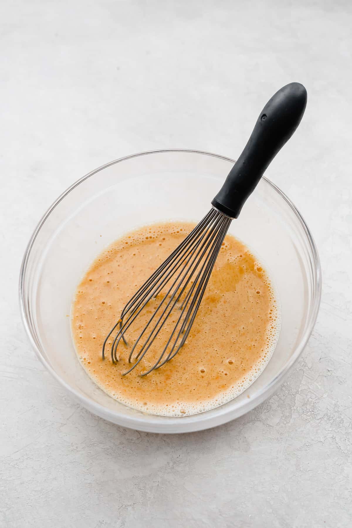 A whisk stirring a light orange liquid mixture in a glass bowl.