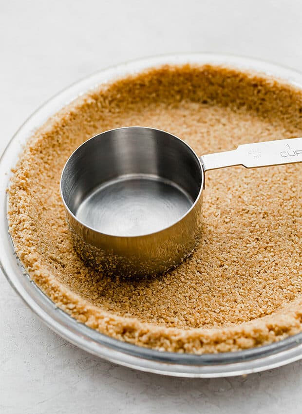 A metal measuring cup pushing down the graham cracker crust.