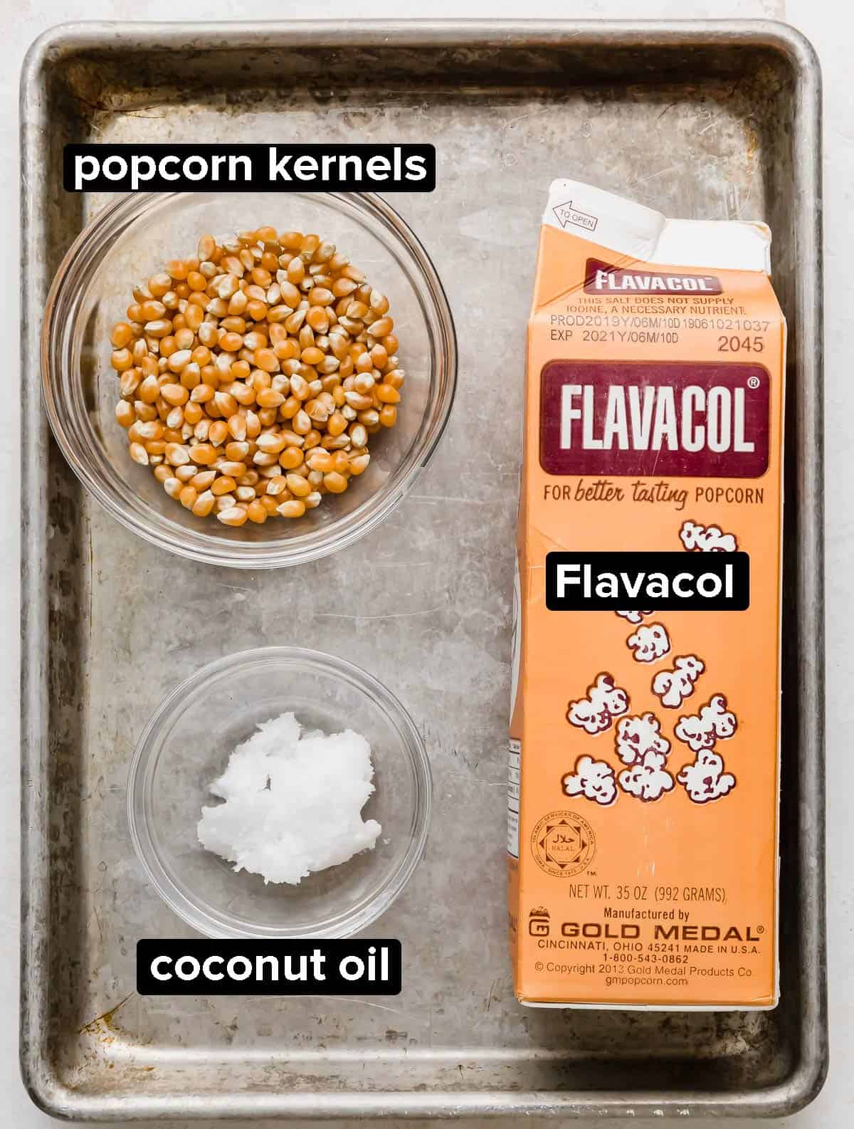 Popcorn kernels, coconut oil, and Flavacol on a gray baking sheet.