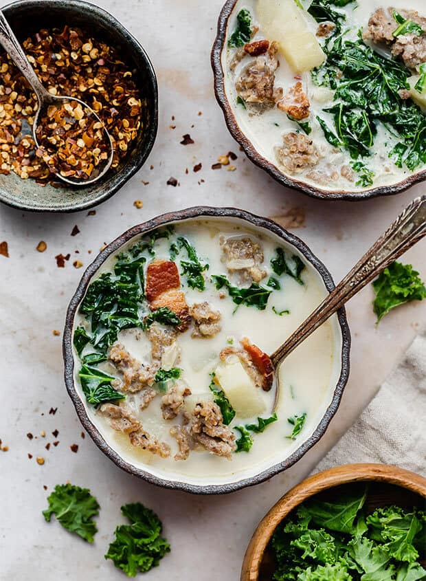 A bowl of Zuppa Toscana with a bowl of kale and crushed pepper flakes nearby.