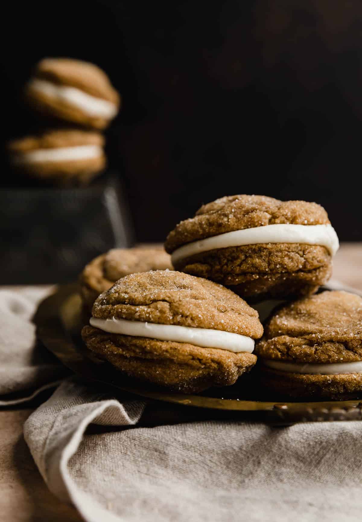 Gingerbread Whoopie pies with a cream cheese filling, stacked on a bronze plate against a brown background.