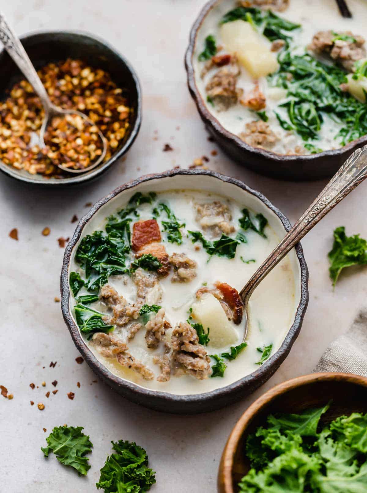 Olive Garden Zuppa Toscana Soup recipe in a black rimmed bowl on a cream textured background with a black bowl filled with crushed red pepper flakes in the background.