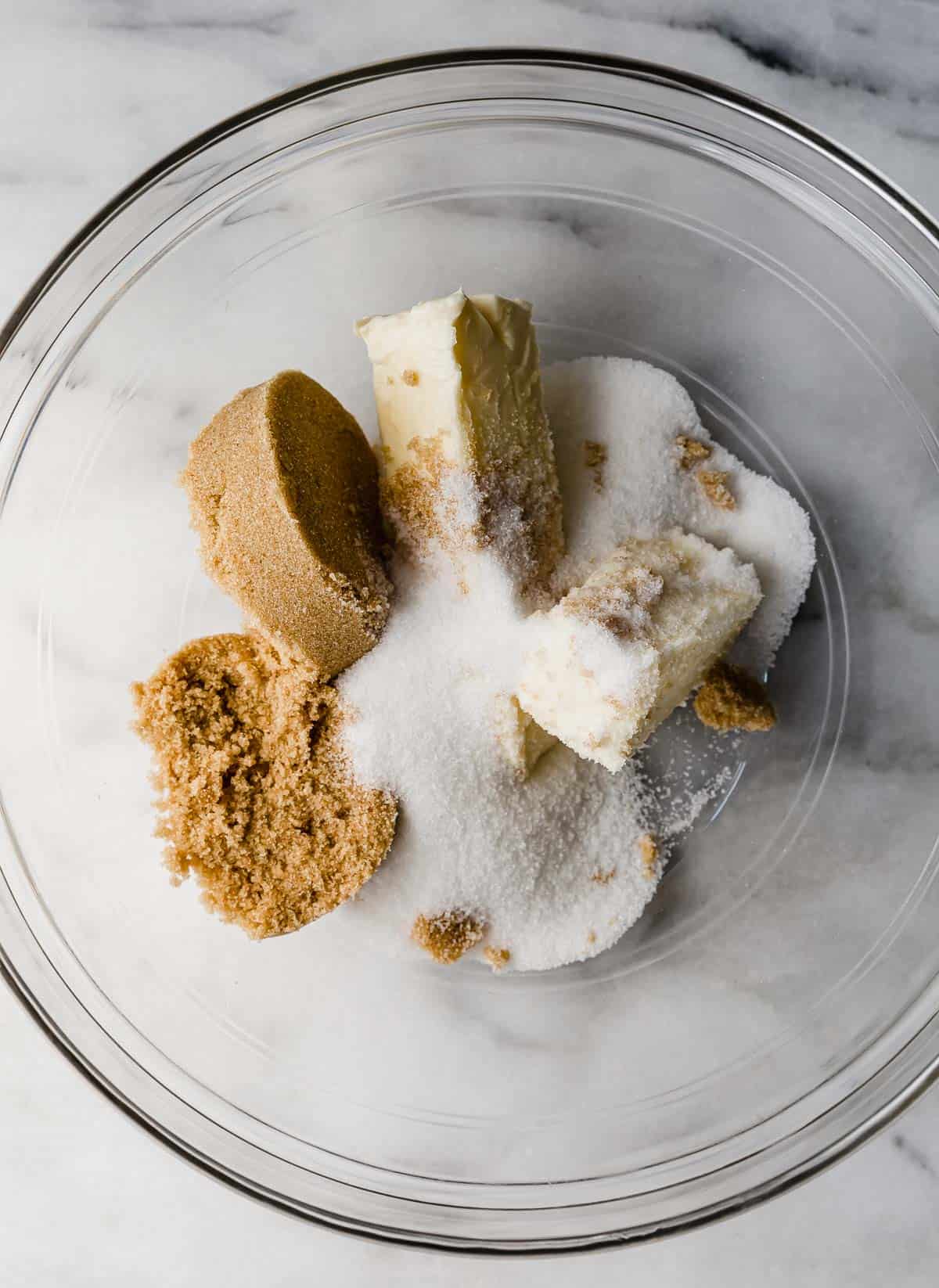 Butter, sugar, and brown sugar in a glass bowl on a marble background.