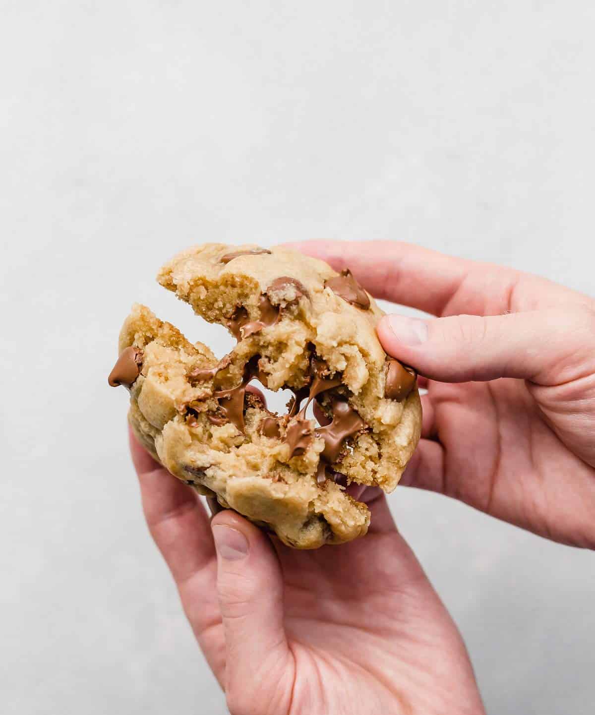 A pair of male hands holding a Crumbl Chocolate Chip Cookie and splitting it in half.