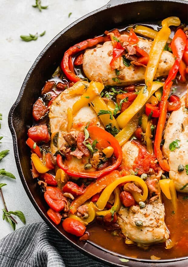 A skillet with cooked Roman chicken topped with fresh herbs, capers, and sliced peppers.