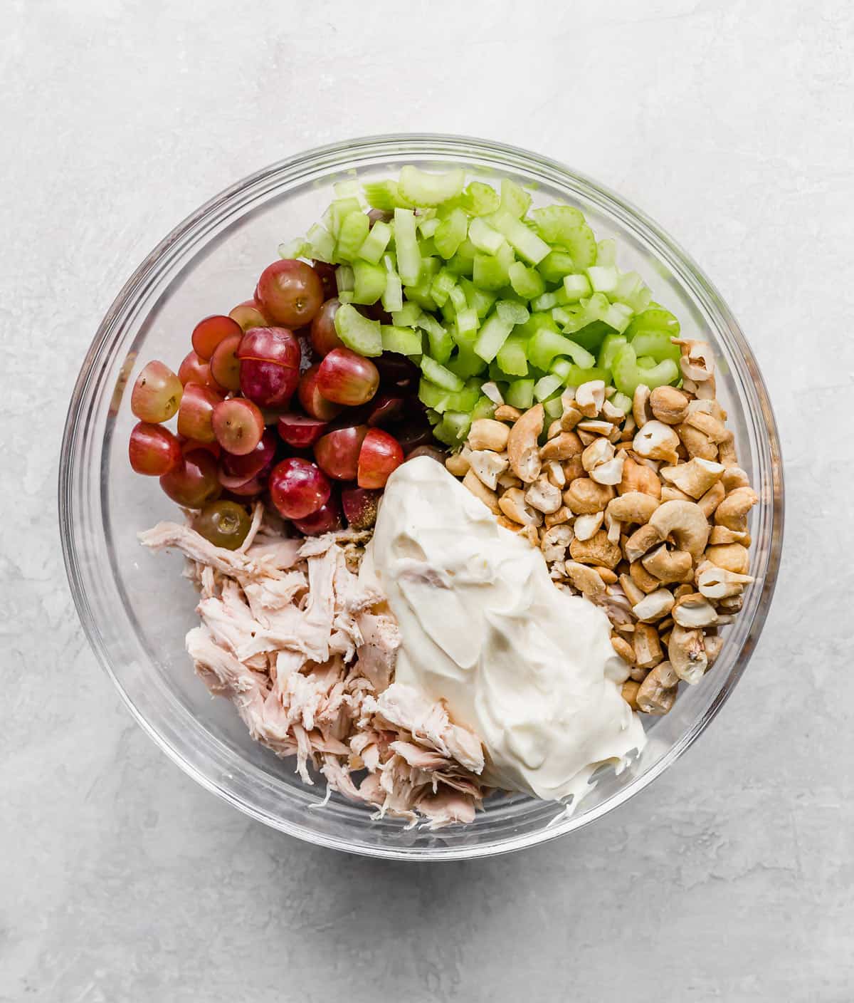 A glass bowl with shredded chicken, purple grapes, celery, cashews, and mayo in it.
