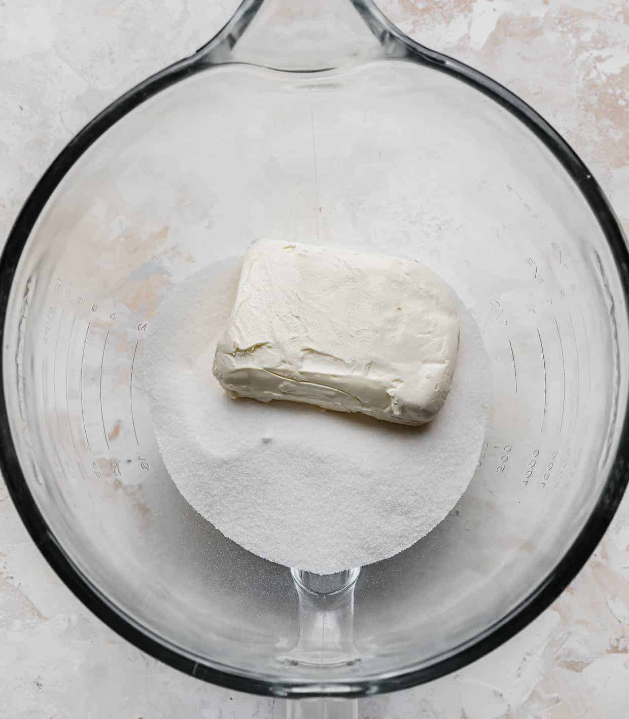 A glass mixing bowl with granulated sugar and a block of white cream cheese in it.