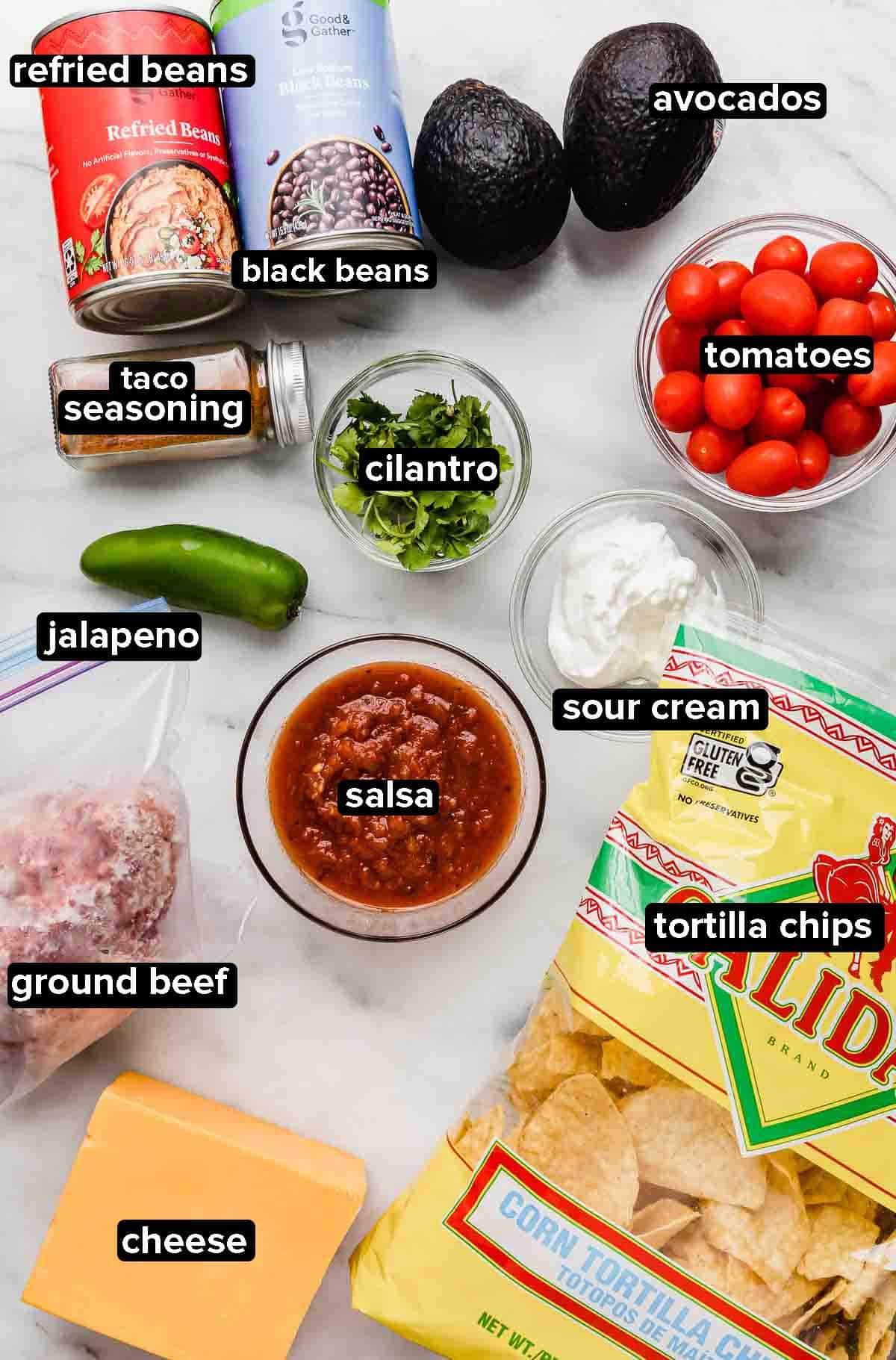 Beef Loaded Nachos ingredients on a white background including: tortilla chips, ground beef, tomatoes, jalapeño, refried beans, avocado, black beans, tomatoes, and cheese.