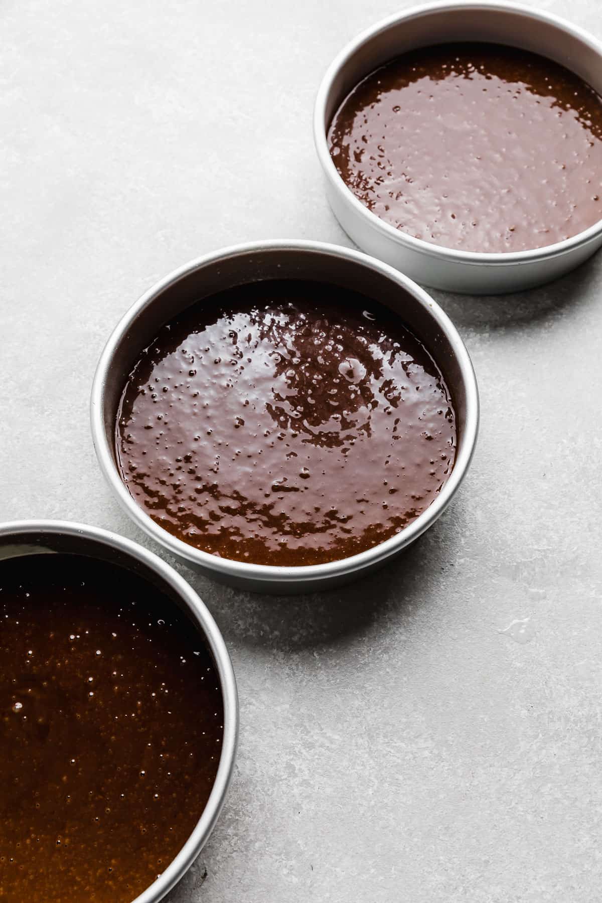 Three round cake pans filled with dark chocolate cake batter made with oil.