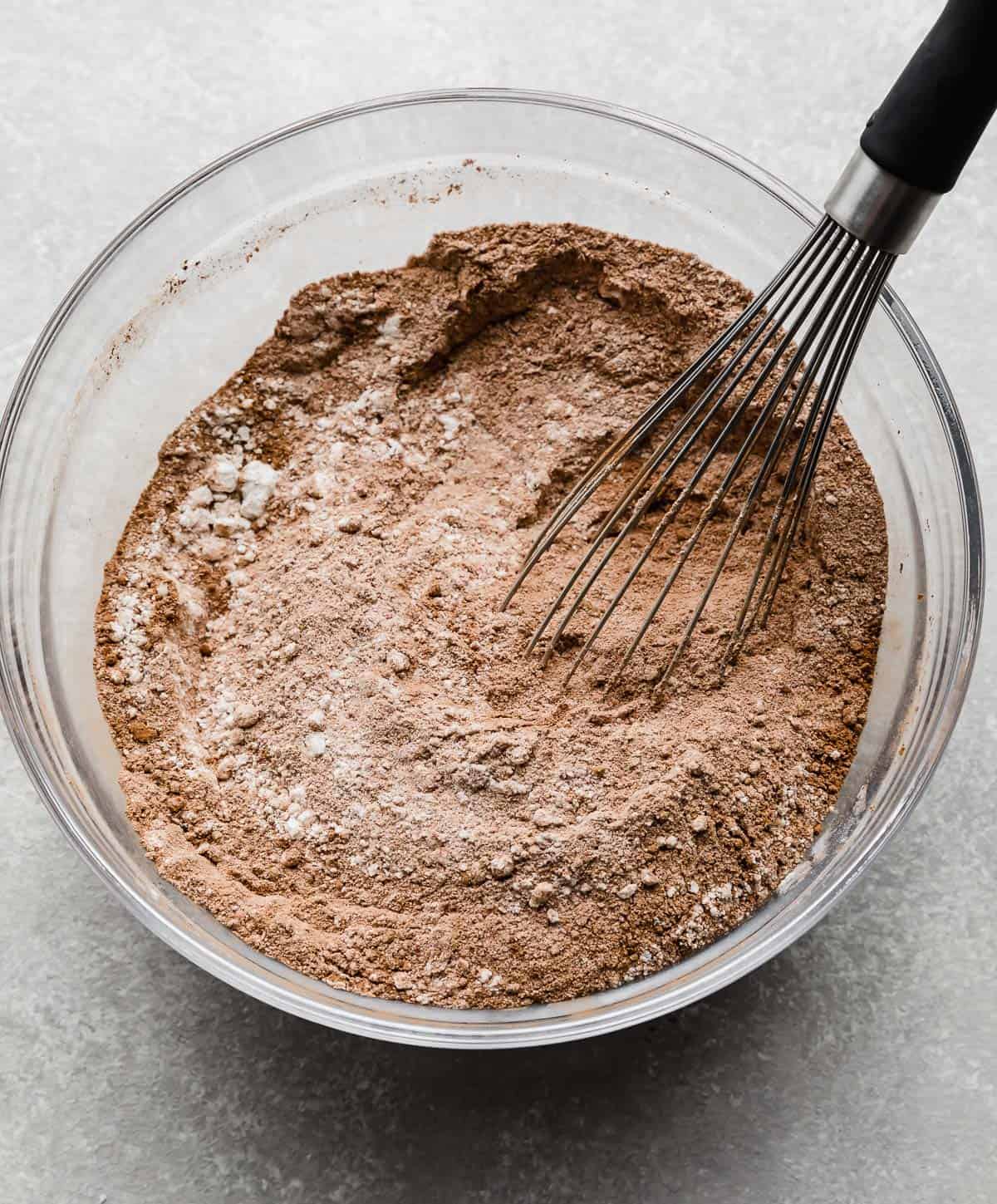 A whisk stirring brown dry ingredients in a glass bowl.