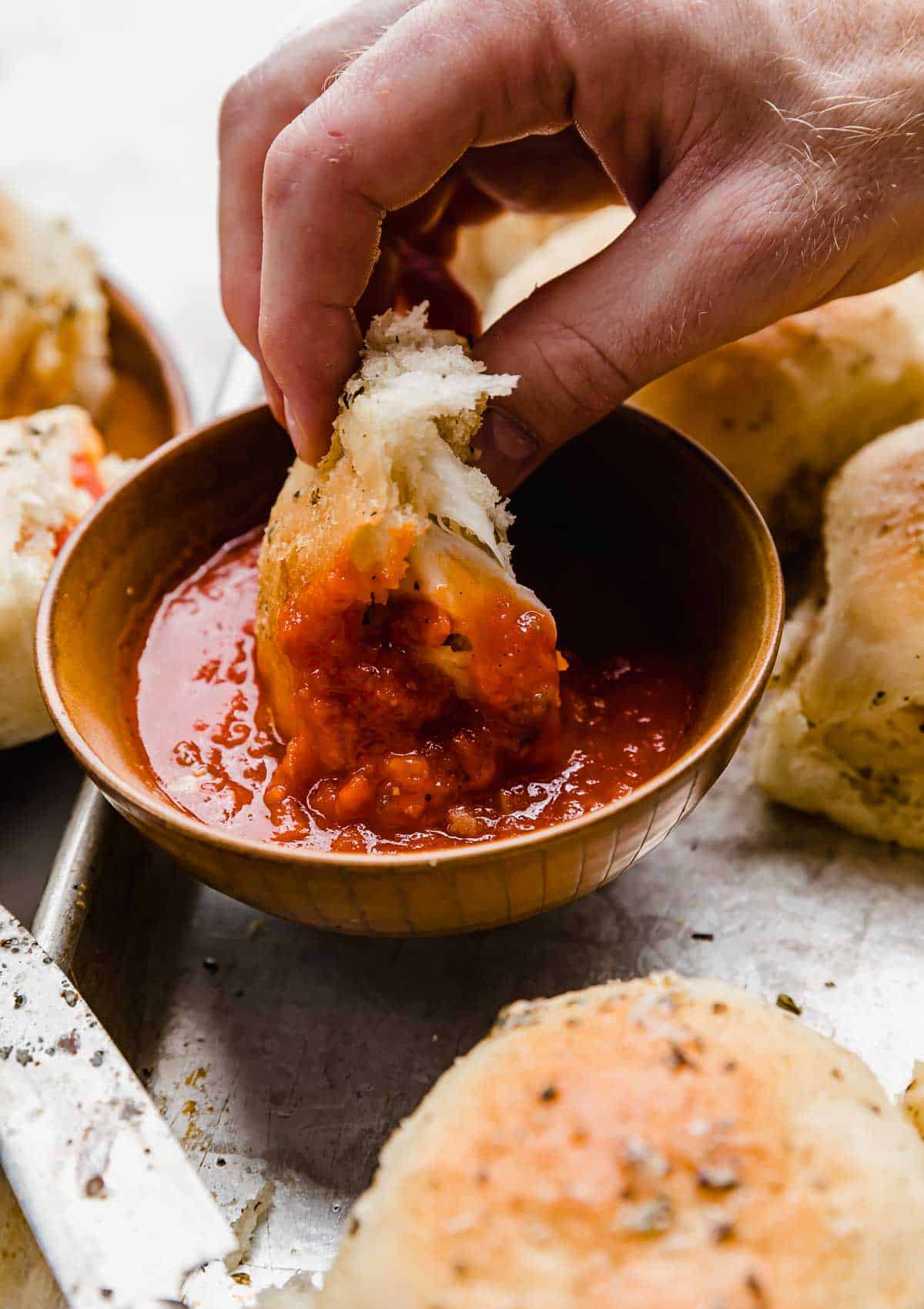 A hand dipping a pepperoni pizza roll in red pizza sauce.