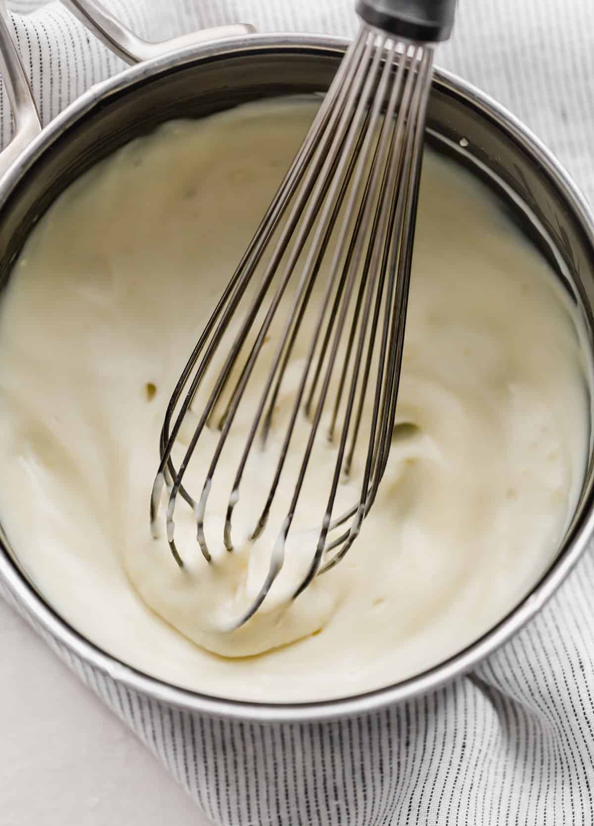 A white thick mixture in a pan with a whisk stirring.
