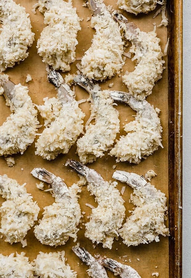 Raw shrimp dipped in a batter and coconut bread crumb mixture, sitting on a baking sheet.