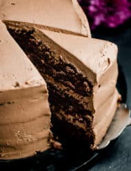 A slice of a three layer Moist Chocolate Cake topped with chocolate buttercream on a black background.