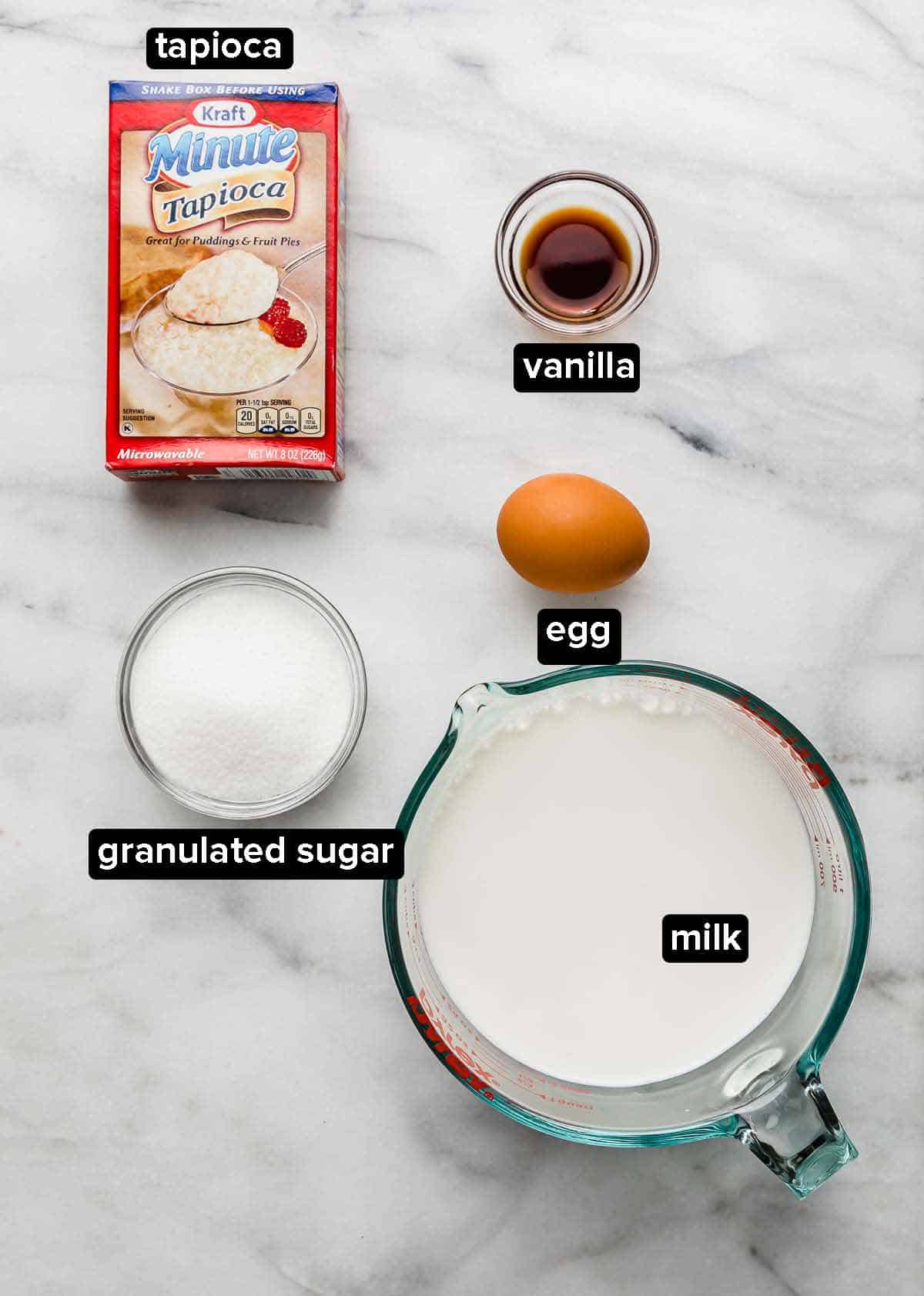 Tapioca Pudding Recipe ingredients on a white marble background: instant tapioca, milk, sugar, egg, and vanilla.
