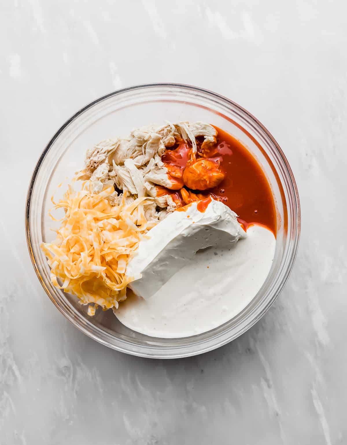 A glass bowl with shredded chicken, cream cheese, buffalo sauce, ranch dressing, and cheese in it.