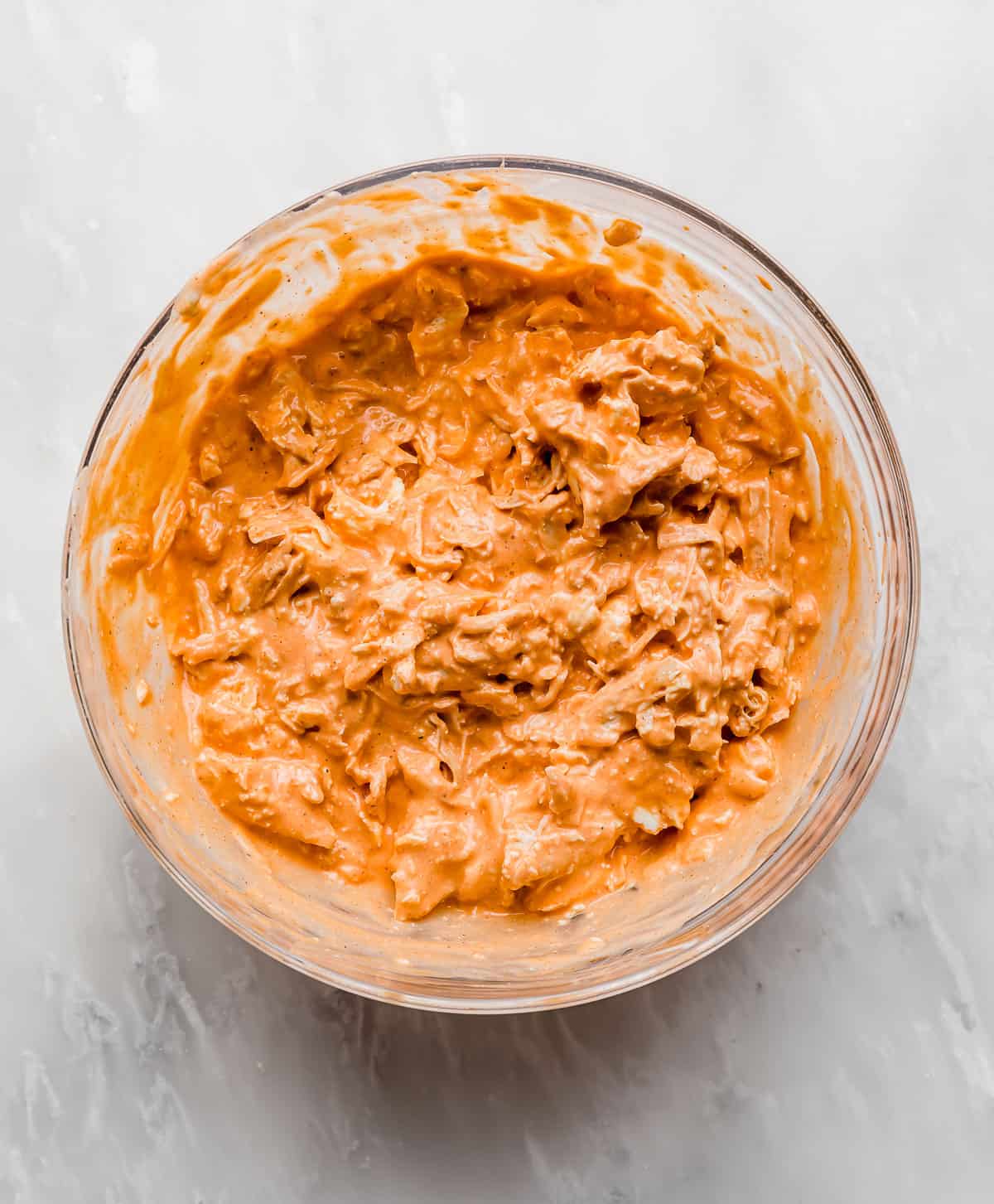 Bright orange Buffalo Chicken Dip mixture in a glass bowl on a white marble background.