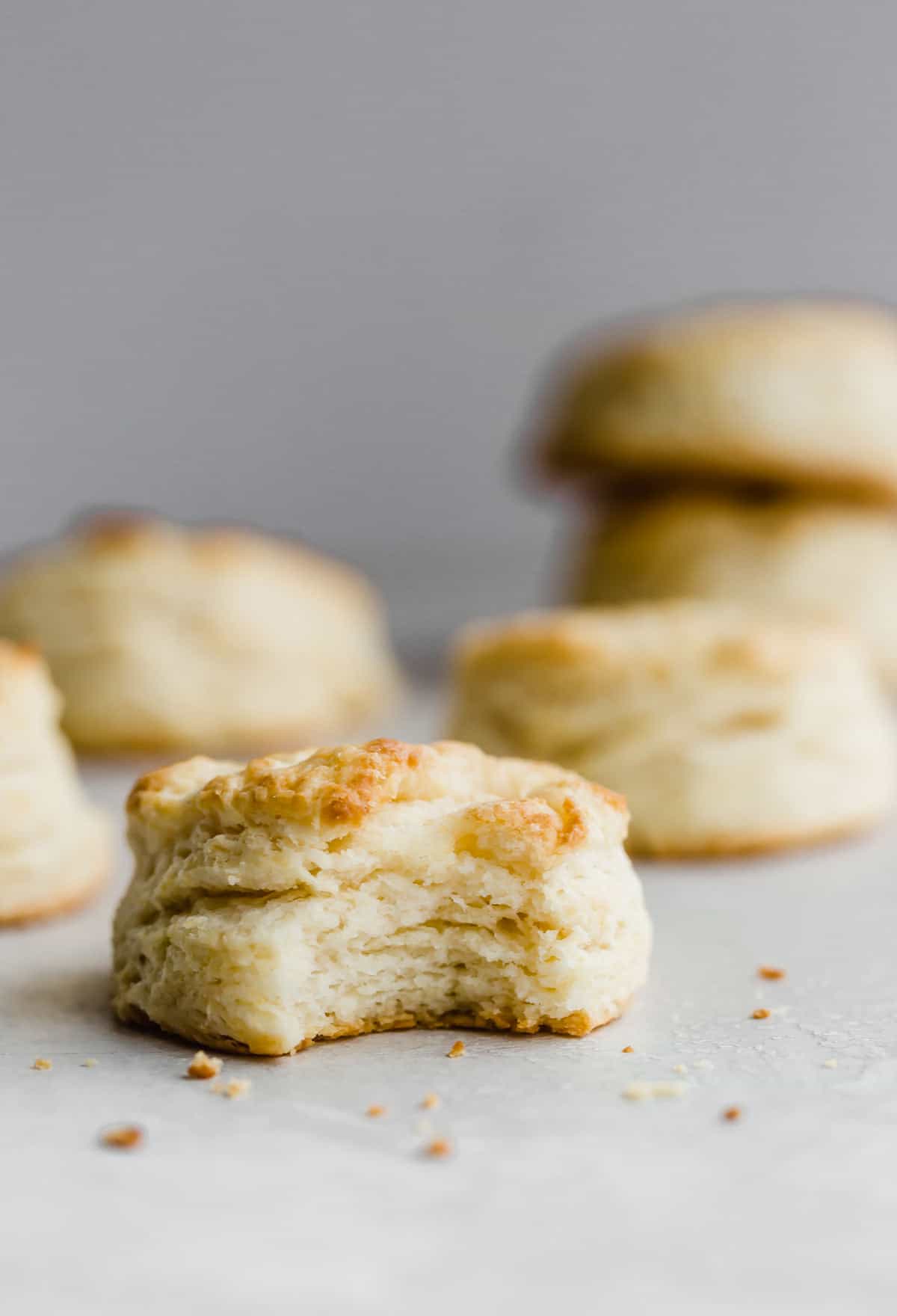 A flaky buttermilk biscuit with a bite taken out of it, against a light gray background. 