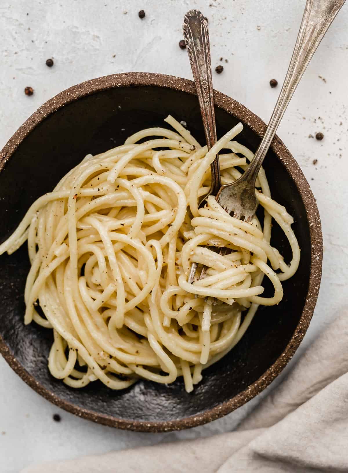 A black plate with two forks twirled into Bucatini Cacio e Pepe noodles.