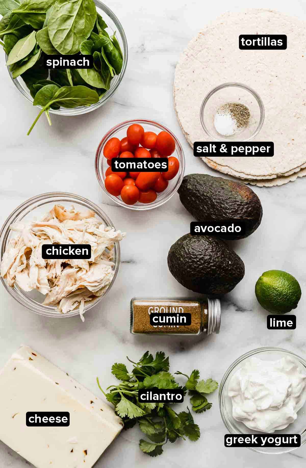 Healthy Chicken Wraps ingredients portioned into glass bowls on a white background: tortillas, spinach, tomatoes, chicken, avocado, lime, cheese, greek yogurt.