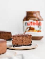 A slice of Nutella Cheesecake on a plate with a Nutella jar in the background.