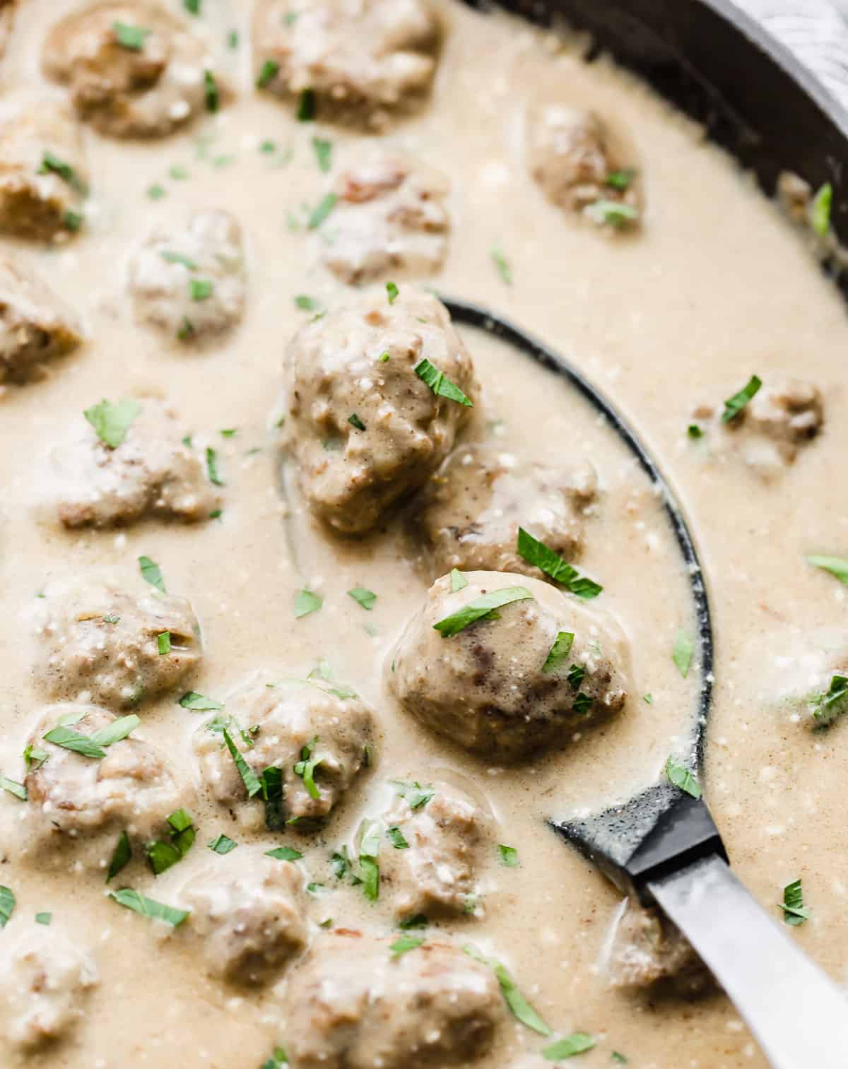 A large spoon scooping into a skillet full of Swedish Meatballs and tan colored sauce.