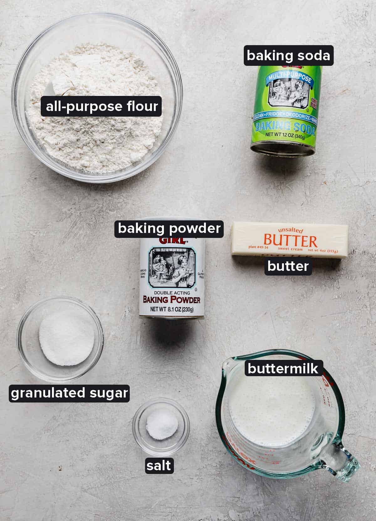 Buttermilk biscuit ingredients portioned into glass bowls on a light gray textured background: flour, buttermilk, baking powder and soda, salt, sugar, and butter.