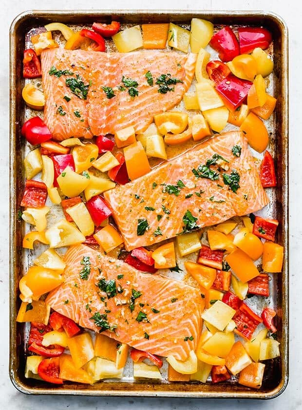 Overhead photo of 3 salmon pieces on a baking sheet surrounded by yellow, orange, and red bell pepper pieces.