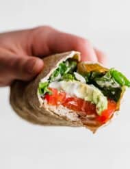 A hand holding a Healthy Chicken Wrap with a bite taken out of the wrap showing the avocado, tomato, chicken, and spinach in the wrap.