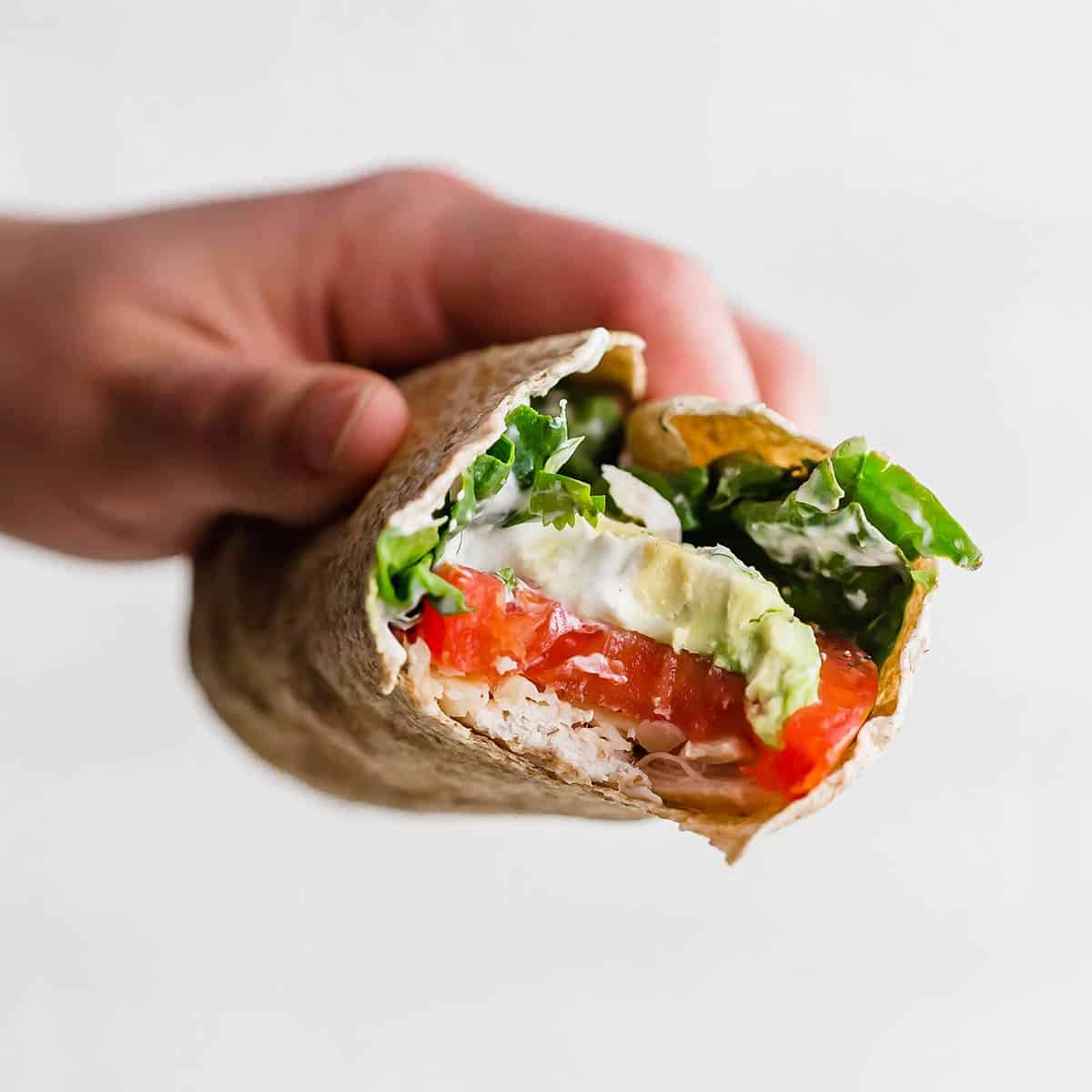 A hand holding a Healthy Chicken Wrap with a bite taken out of the wrap showing the avocado, tomato, chicken, and spinach in the wrap.