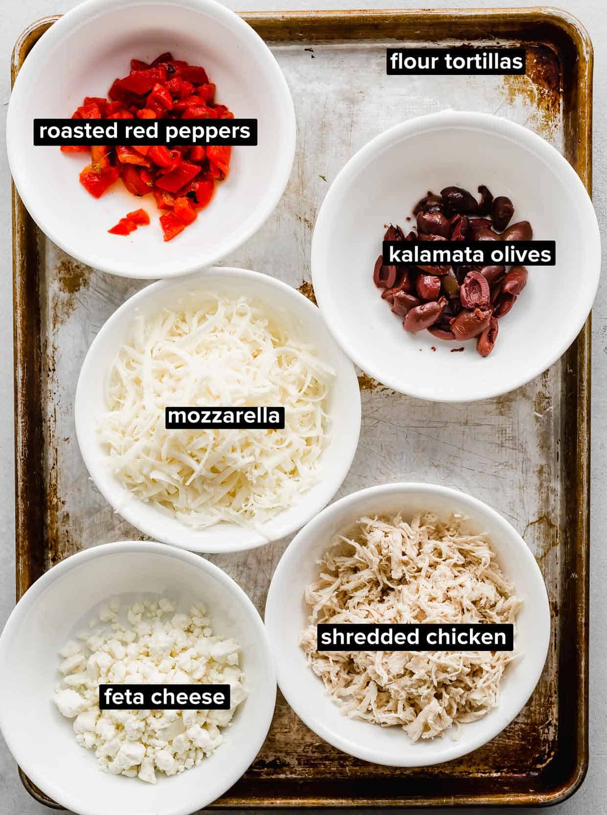 Ingredients used to make Greek Quesadillas portioned into white bowls on a seasoned baking sheet.