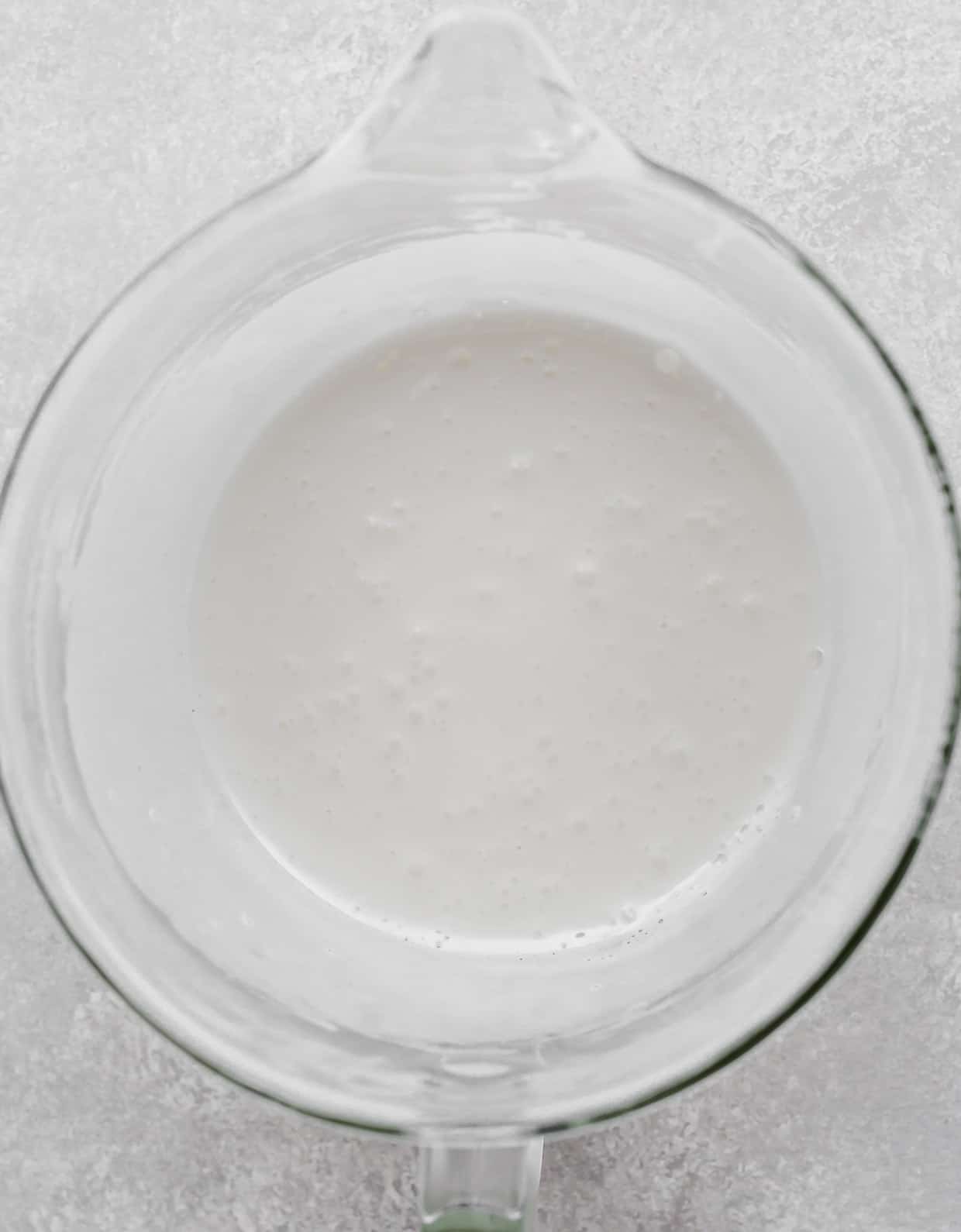 A glass stand mixer bowl with a white liquid mixture in it on a light gray background.