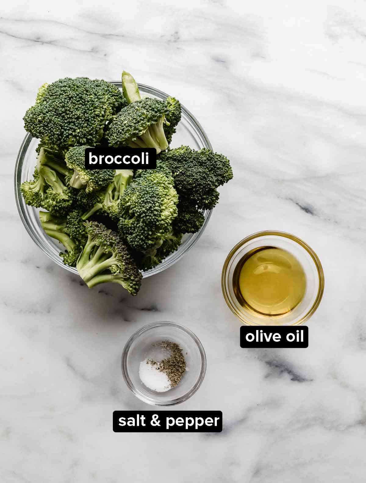 Oven Roasted Broccoli ingredients on a white marble background: broccoli florets, olive oil, salt and pepper.