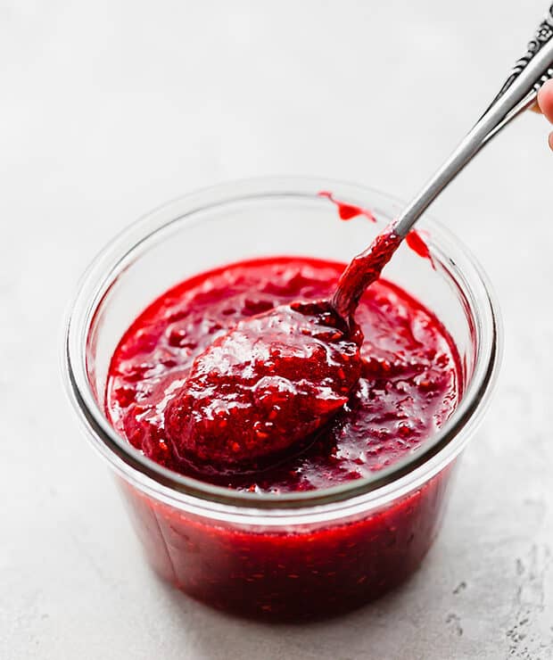 A spoon scooping out raspberry freezer jam from a glass jar.