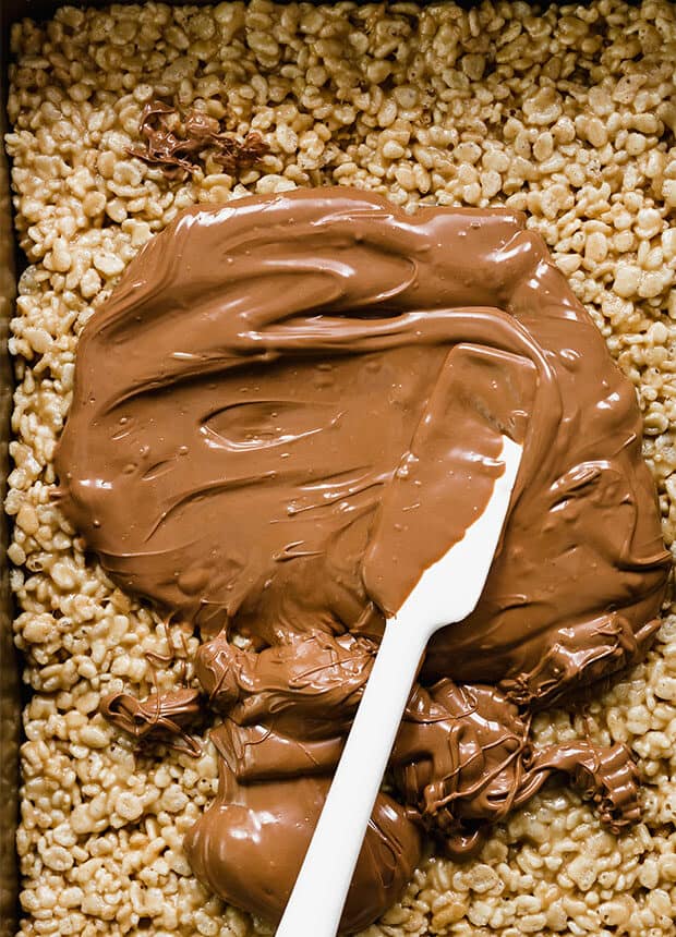 A spatula spreading melted chocolate over scotcharoo Rice Krispies.