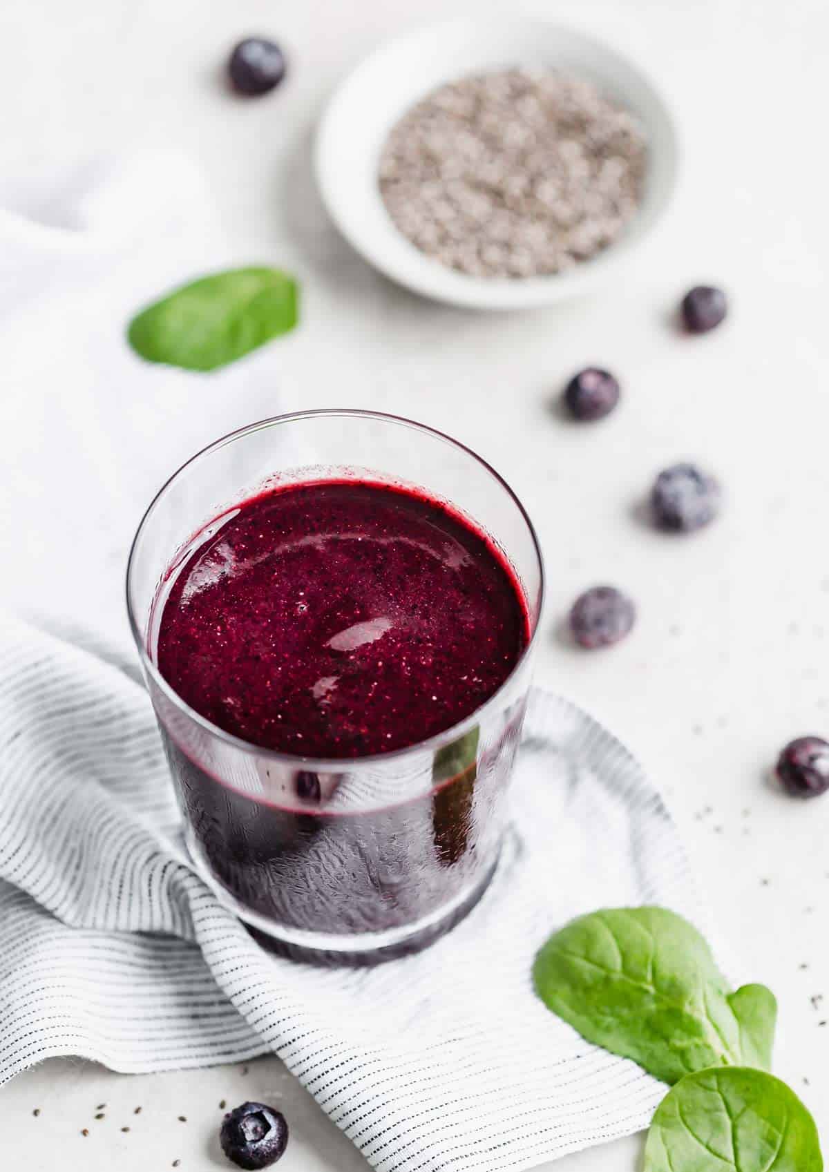 A clear cup filled with a deep purple Acai Smoothie on a white background with frozen blueberries scattered around the cup.