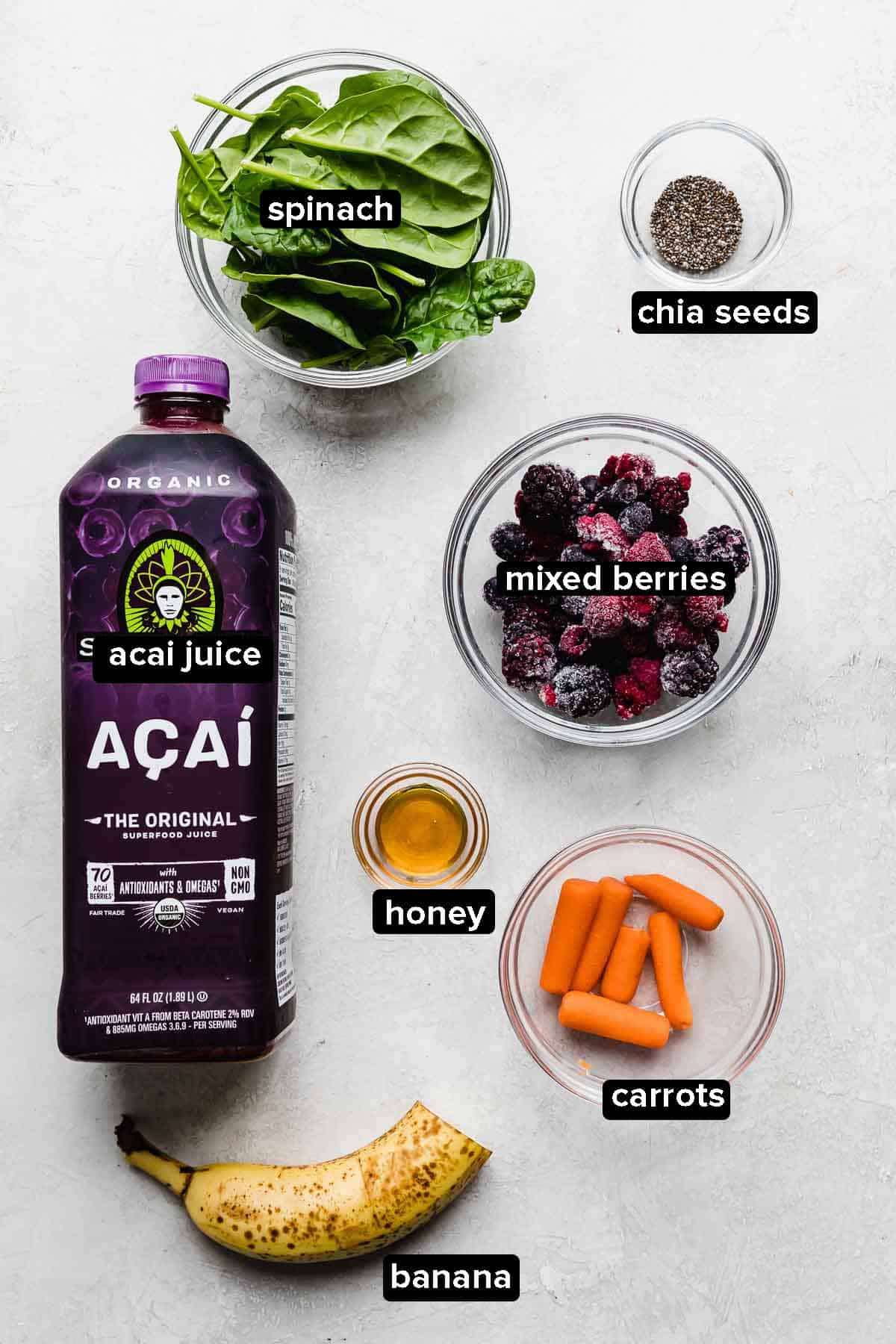 Acai Smoothie ingredients on a white background: carrots, mixed berries, spinach, açaí juice, chia seeds, and honey.