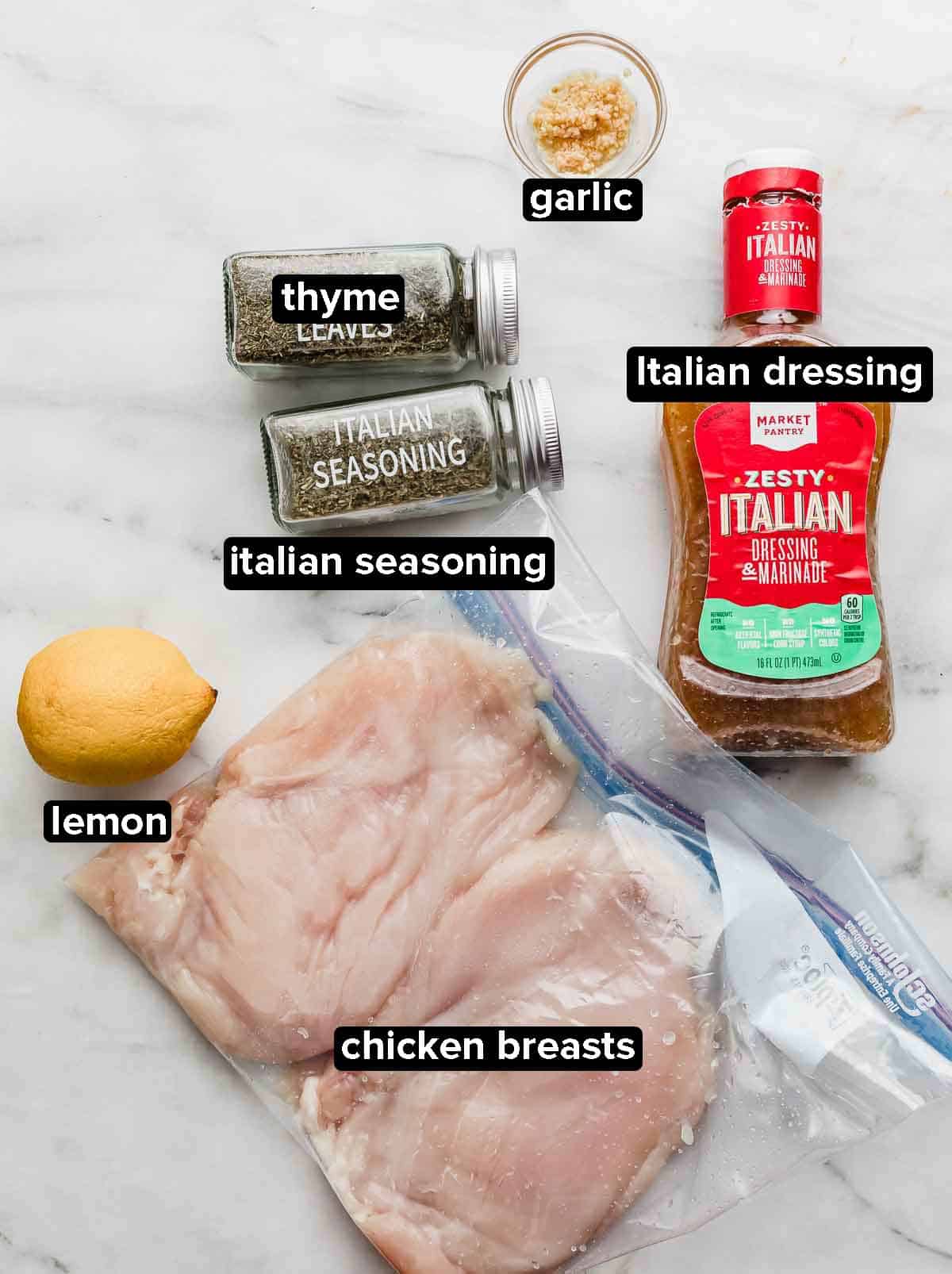 Italian Grilled Chicken ingredients on a white background: raw chicken breasts, lemon, Italian salad dressing, garlic, thyme and Italian seasoning.
