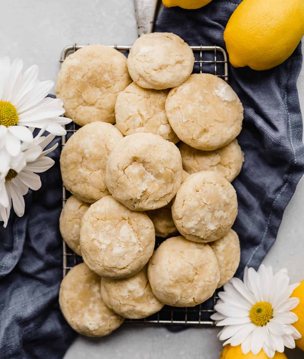 A stack of Lemon Cookies on a small wire rack that is resting on a dark blue linen napkin with two daisy flowers near the cookies.