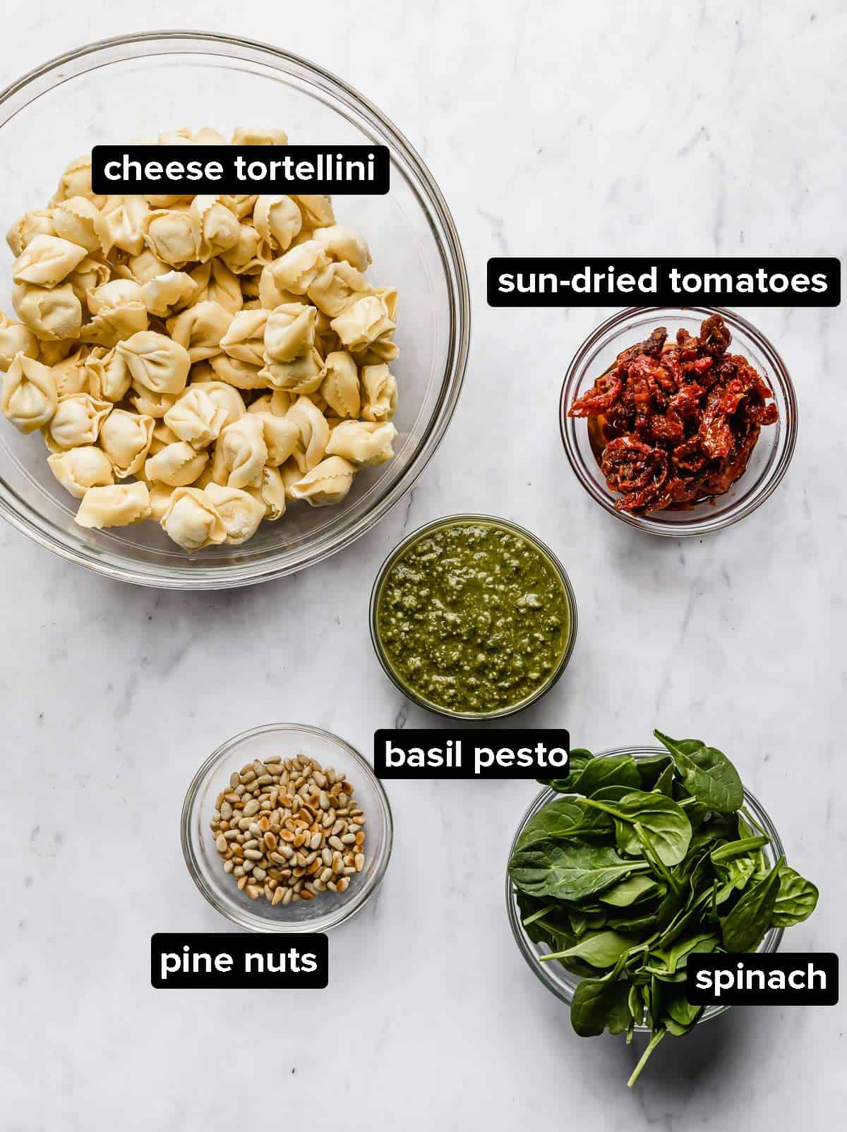 Ingredients used to make Pesto Tortellini Pasta Salad, portioned into glass bowls: tortellini, pesto, sun-dried tomatoes, pine nuts, and optional spinach.