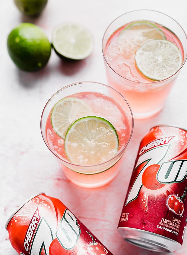 A glass of limeade slush with limes and cherry 7-UP surrounding it.