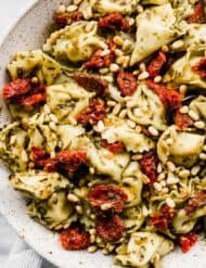 A close up photo of Pesto Tortellini Pasta Salad tossed with sun-dried tomatoes and pine nuts in a white bowl.