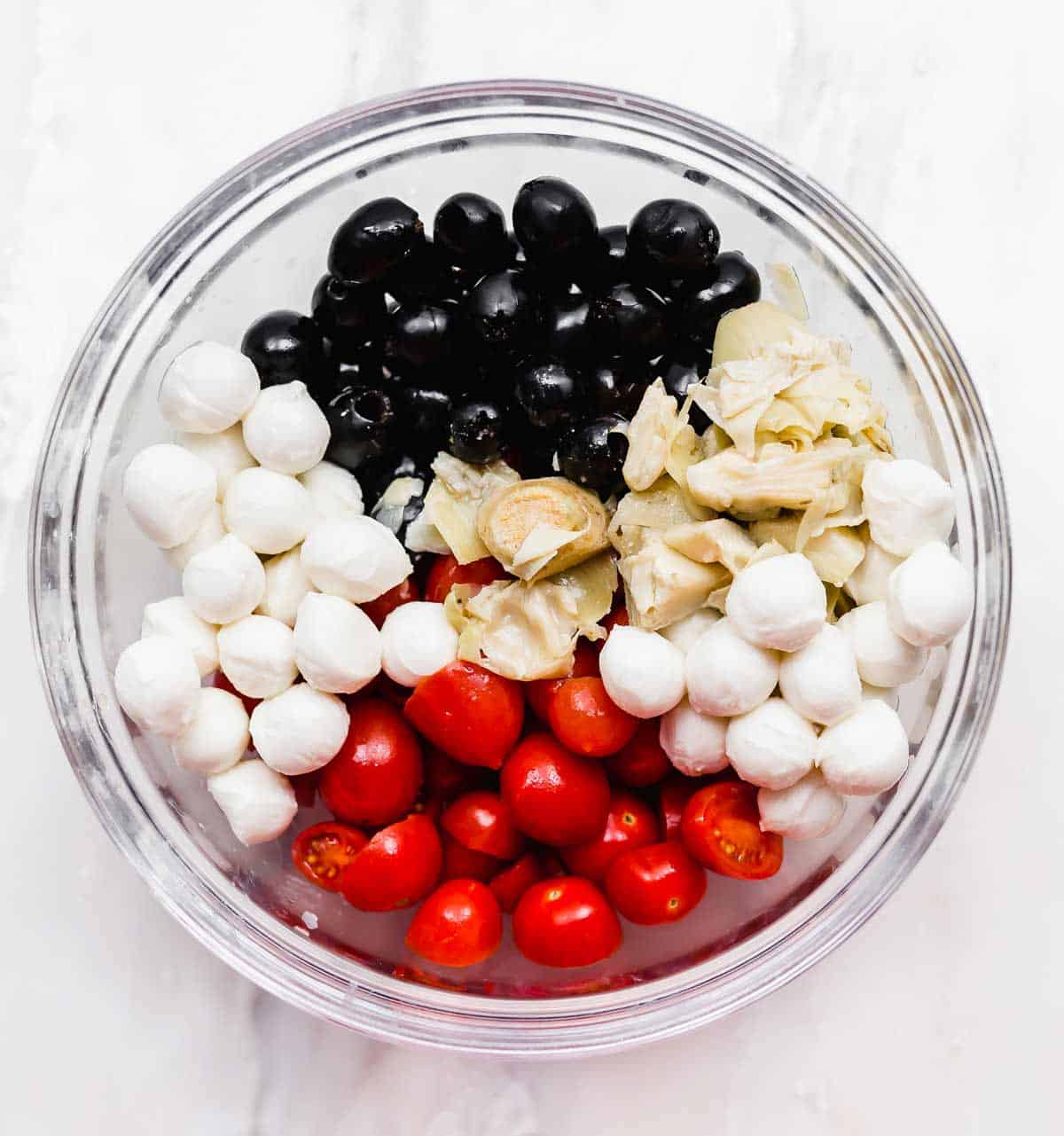 A glass bowl filled with mozzarella pearls, tomatoes, olives, and artichokes.